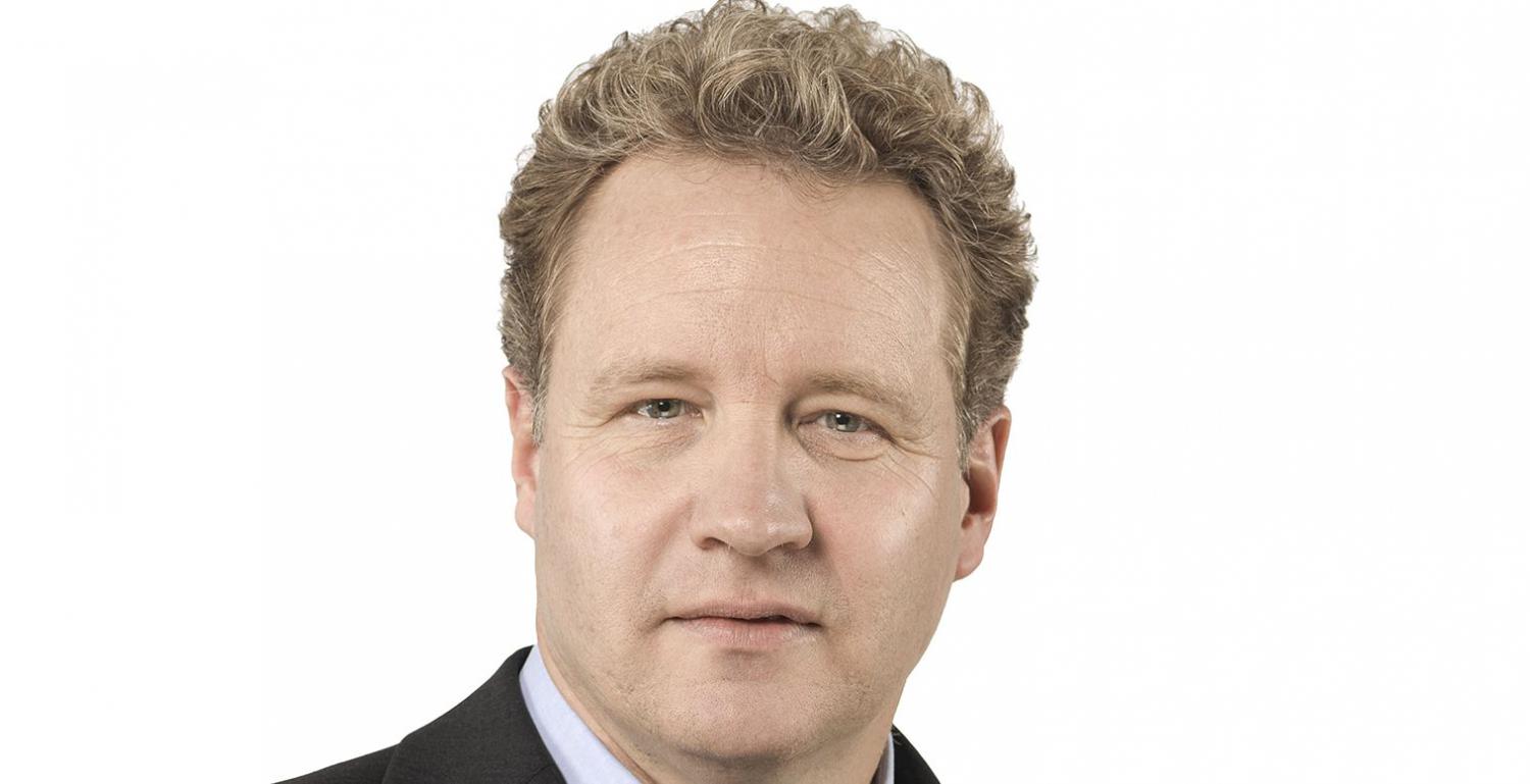 Porträt eines Mannes mit lockigem, blondem Haar, in einem dunklen Anzug, vor hellem Hintergrund.