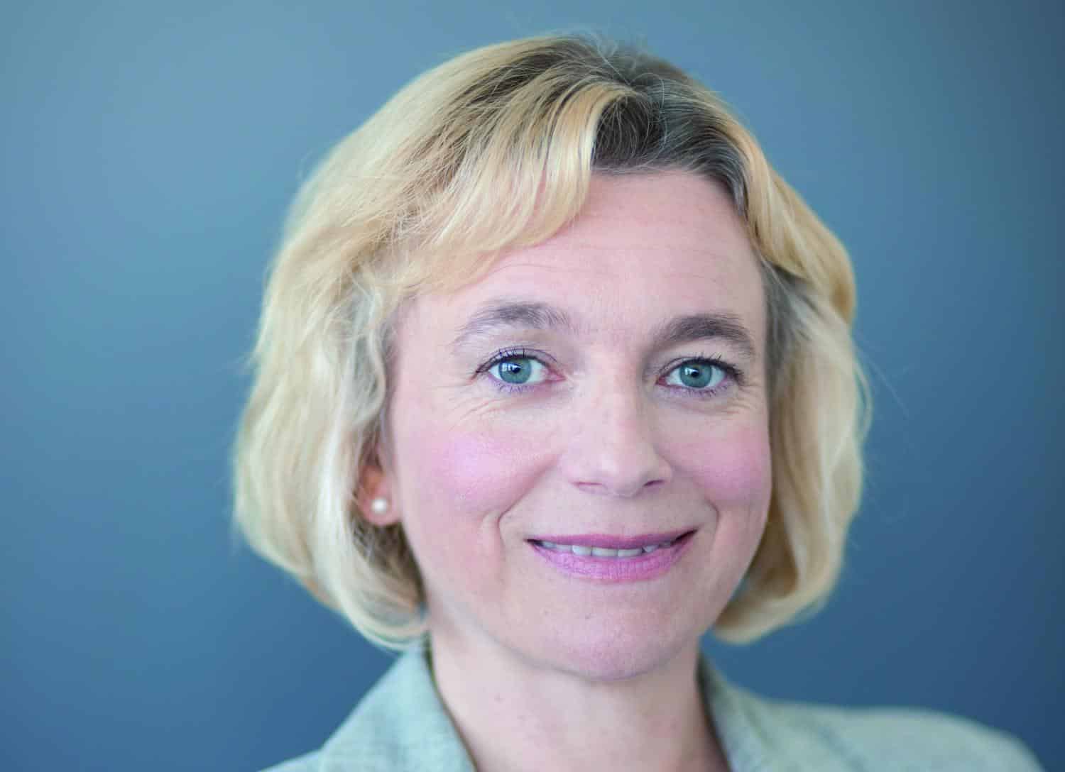 Porträt einer Frau mit blonden, lockigen Haaren und blauen Augen, lächelnd vor einem grauen Hintergrund.