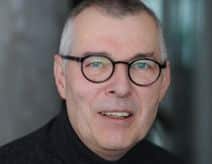 Porträt eines Mannes mit Brille, lächelnd, vor unscharfem Hintergrund.