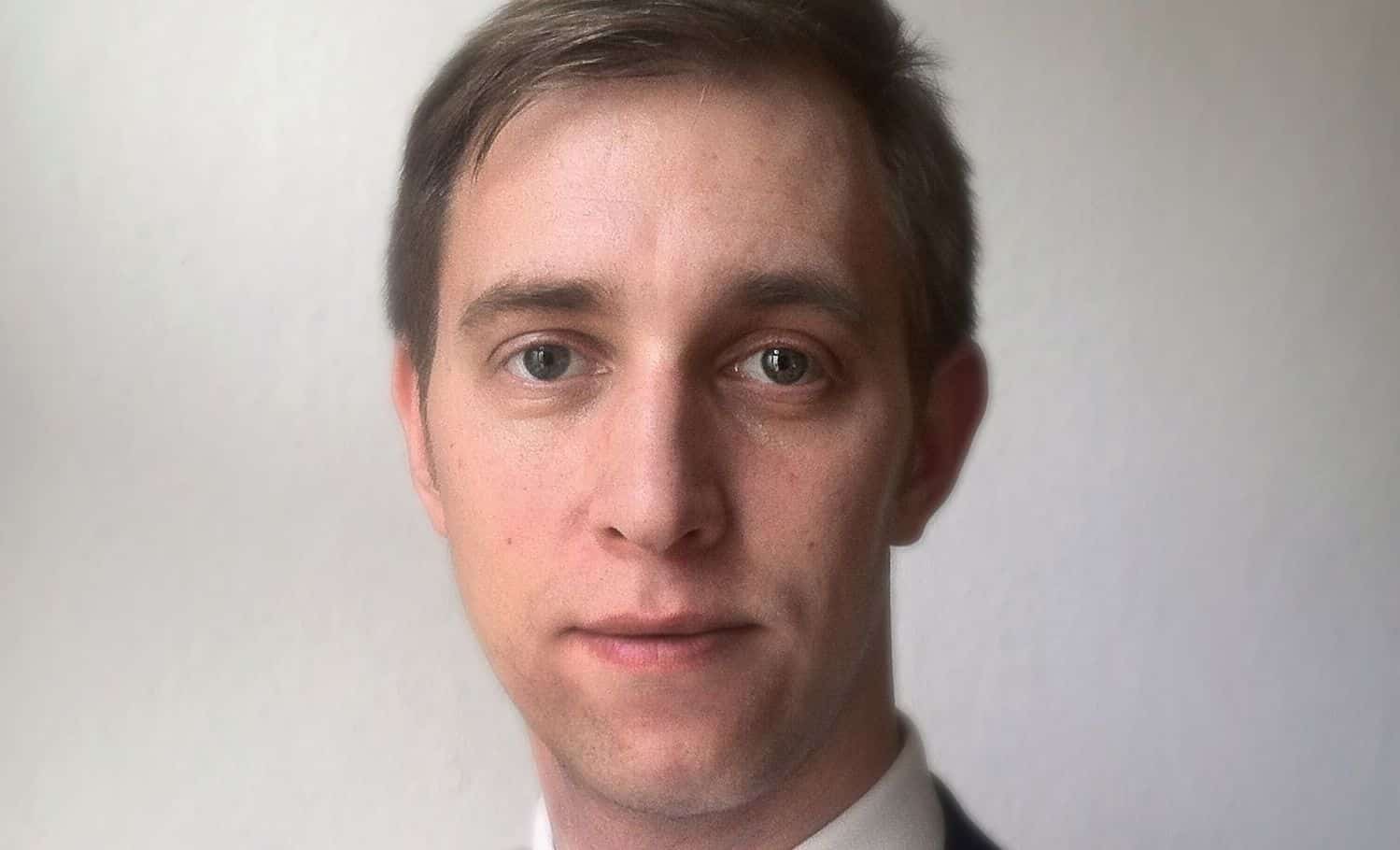 Porträt eines Mannes mit kurzen, braunen Haaren und einem neutralen Gesichtsausdruck vor einem hellen Hintergrund.