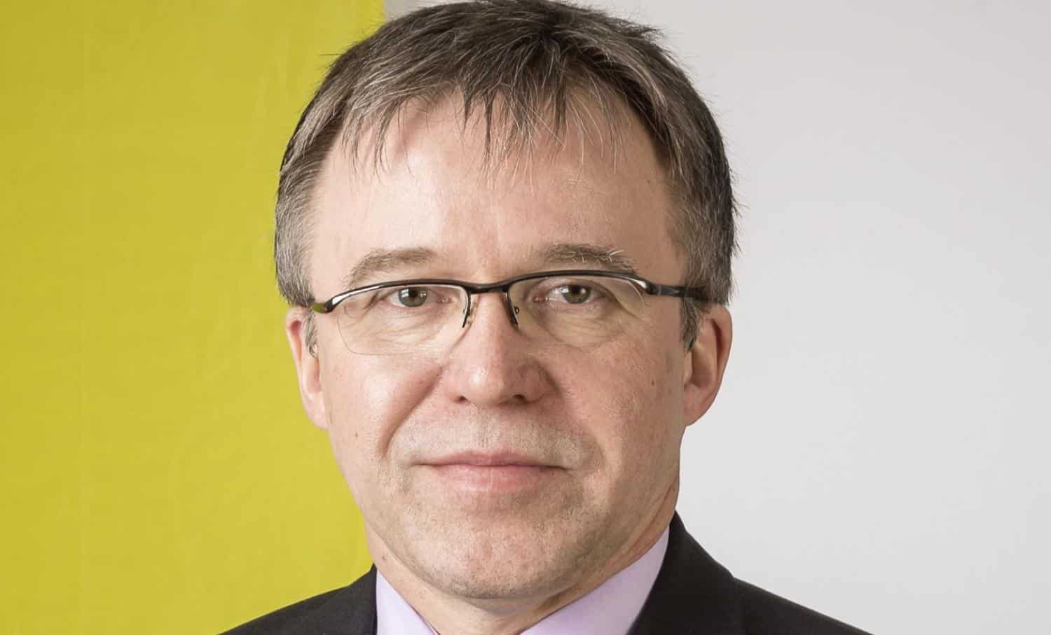 Porträt eines Mannes mit Brille, grauen Haaren und Anzug, vor einem gelben und weißen Hintergrund.