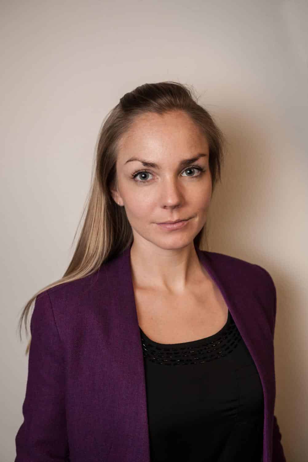 Porträt einer Frau mit langen, blonden Haaren, die einen lila Blazer trägt, vor einem neutralen Hintergrund.