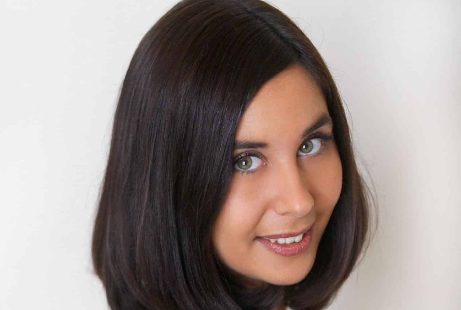 Porträt einer Frau mit schulterlangen, braunen Haaren und blauen Augen, lächelnd vor einem hellen Hintergrund.