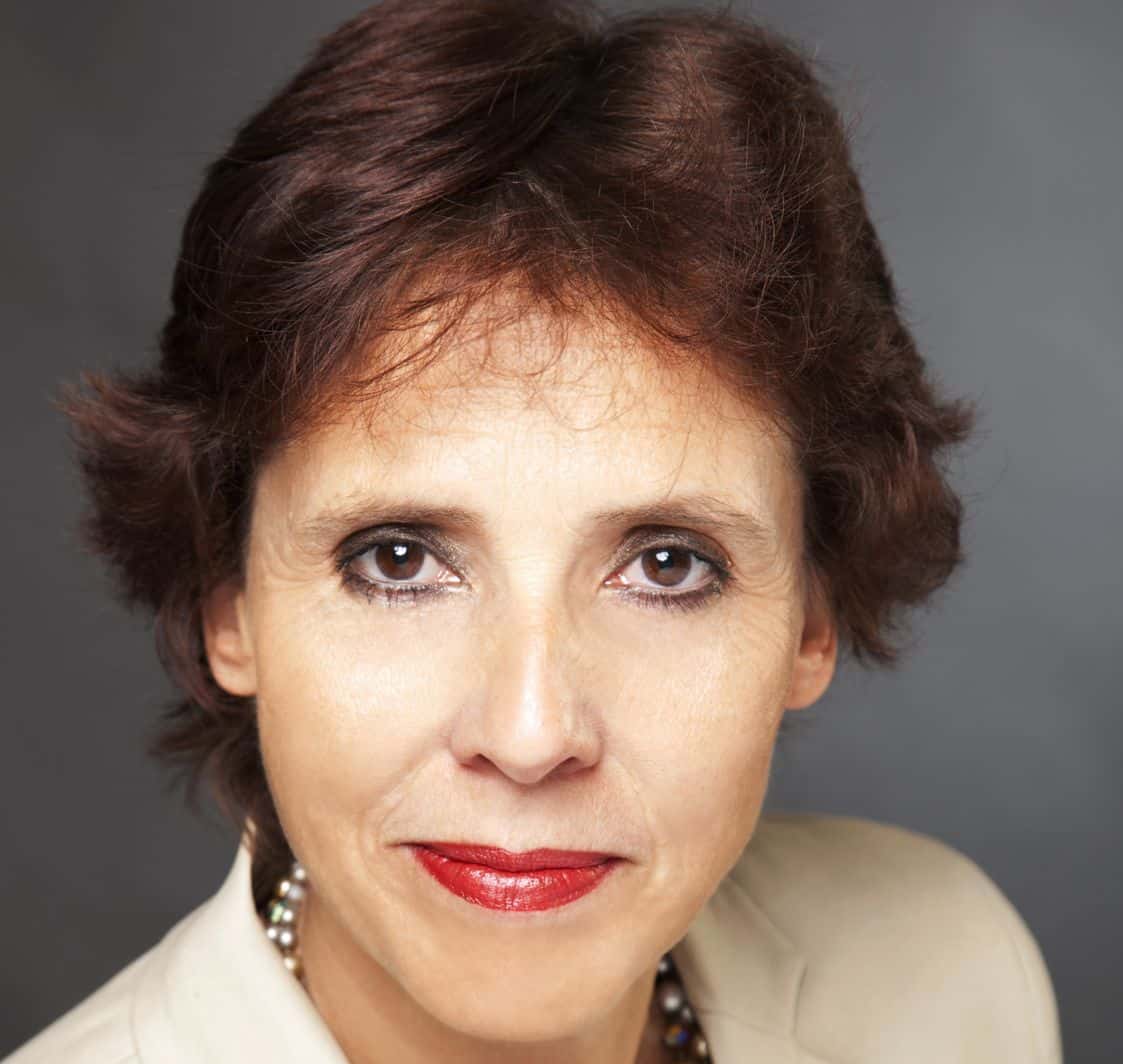 Porträt einer Frau mit kurzen, braunen Haaren, braunen Augen und rotem Lippenstift, vor einem grauen Hintergrund.