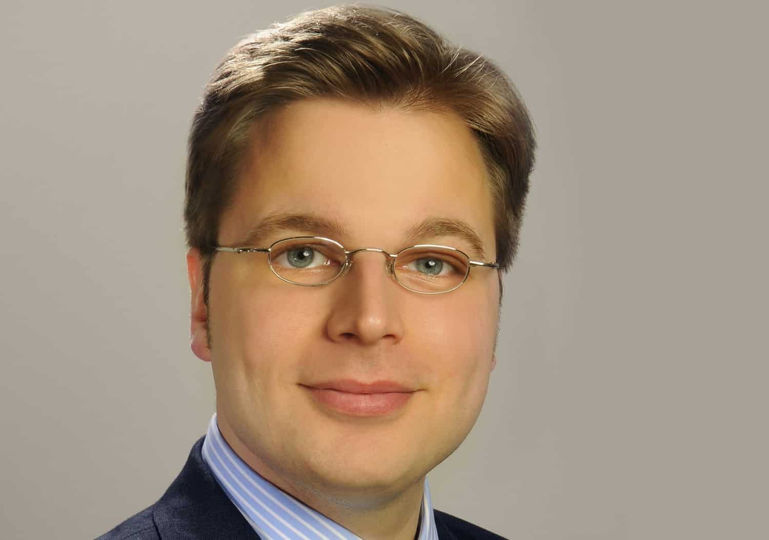 Porträt eines Mannes mit Brille, lächelnd, in Anzug und Krawatte, vor neutralem Hintergrund.