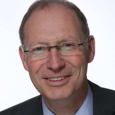 Porträt eines lächelnden Mannes mit Brille, in Anzug und Hemd, vor einem hellen Hintergrund.