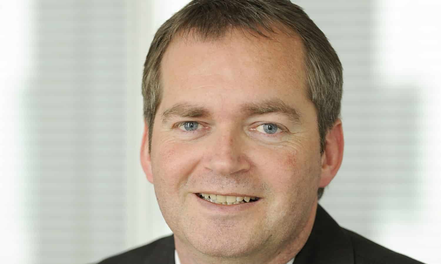 Porträt eines Mannes mit kurzem, grauem Haar, lächelnd, in einem Anzug vor einem hellen, unscharfen Hintergrund.