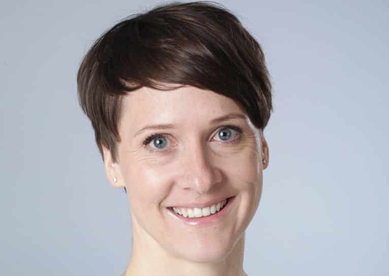 Porträt einer lächelnden Frau mit kurzen, braunen Haaren und blauen Augen vor einem hellen Hintergrund.