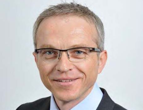 Porträt eines Mannes mit Brille, kurzen Haaren, in einem Anzug, lächelnd vor neutralem Hintergrund.