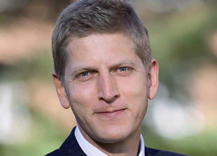 Porträt eines Mannes mit kurzen, blonden Haaren und blauen Augen, lächelnd vor unscharfem Hintergrund.