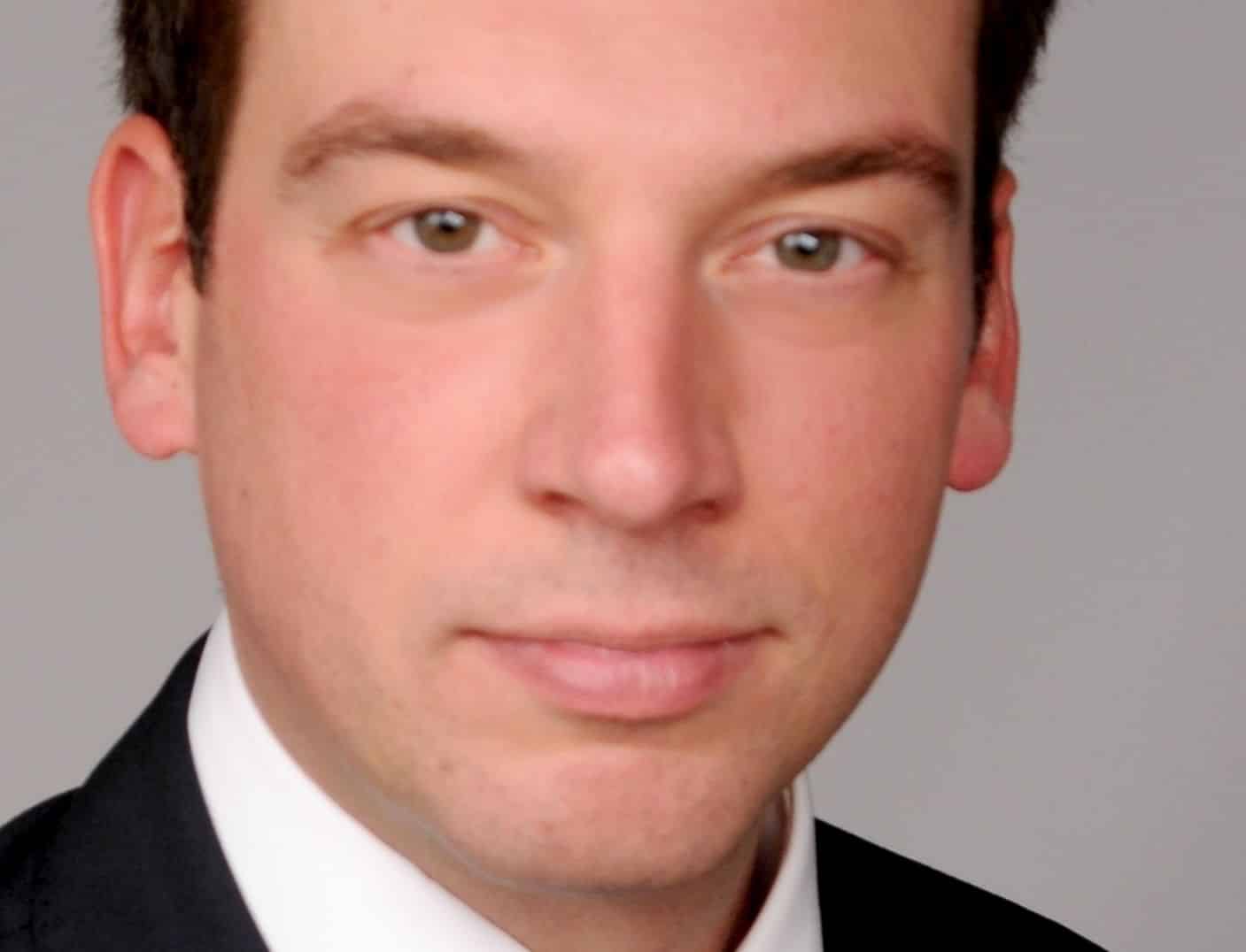 Porträt eines Mannes in Anzug mit kurzen, dunklen Haaren und neutralem Gesichtsausdruck vor grauem Hintergrund.