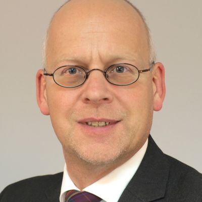 Porträt eines Mannes mit Glatze und Brille, der einen Anzug trägt, vor einem neutralen Hintergrund.