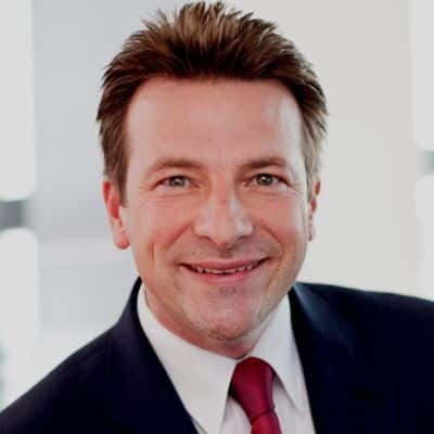 Mann mit braunen Haaren, Anzug und roter Krawatte lächelt vor einem hellen, unscharfen Hintergrund.