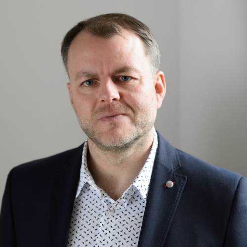 Porträt eines Mannes mit kurzem Haar, in einem dunklen Anzug und einem gemusterten Hemd, vor neutralem Hintergrund.