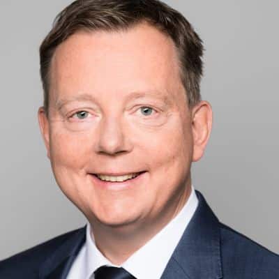 Porträt eines lächelnden Mannes in Anzug mit kurzen, braunen Haaren und blauen Augen vor einem grauen Hintergrund.