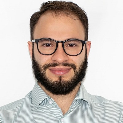 Mann mit Brille und Bart, lächelt vor einem weißen Hintergrund, trägt ein gestreiftes Hemd.