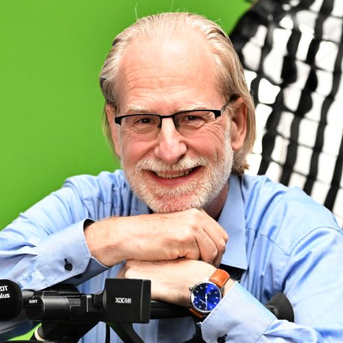 Mann mit Brille und grauen Haaren lächelt, stützt sich auf einen Tisch mit Mikrofon, vor grünem Hintergrund.