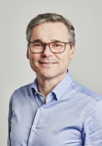 Porträt eines Mannes mit Brille, lächelnd, in einem hellblauen Hemd vor einem neutralen Hintergrund.