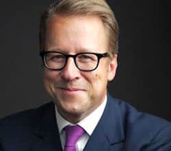 Mann mit Brille und Anzug, lächelt freundlich vor dunklem Hintergrund.