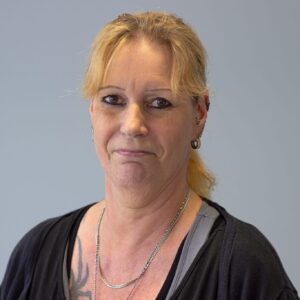 Porträt einer Frau mit blonden Haaren, die einen neutralen Gesichtsausdruck hat, vor einem grauen Hintergrund.