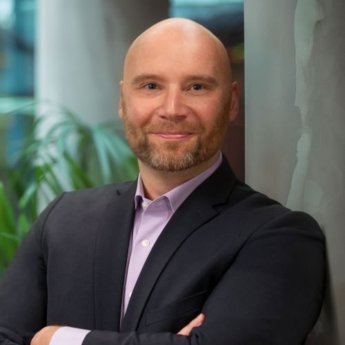 Mann mit Glatze und Bart, in Anzug, lächelt und lehnt an einer Wand, im Hintergrund Pflanzen.