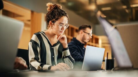 Foto einer Frau mit Brille und hochgesteckten braunen lockigen Haaren, die zwischen zwei Kollegen vor ihrem Laptop sitzt. Dazu die Bildunterschrift: Beschäftigte in der Kommunikationsbranche zeigen sich besonders kritisch, wenn es um die Kommunikation ihres Unternehmens geht. © gorodenkoff/Getty Images