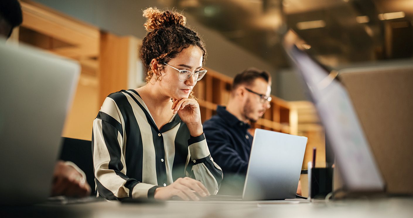 Foto einer Frau mit Brille und hochgesteckten braunen lockigen Haaren, die zwischen zwei Kollegen vor ihrem Laptop sitzt. Dazu die Bildunterschrift: Beschäftigte in der Kommunikationsbranche zeigen sich besonders kritisch, wenn es um die Kommunikation ihres Unternehmens geht. © gorodenkoff/Getty Images