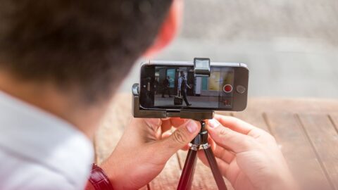 Ein Mann fotografiert an einem Morgen in London mit seinem Smartphone, das auf einem Stativ steht, das auf einem Holztisch platziert ist. Dazu die Bildunterschrift: Für eine professionelle Wirkung ist die Stabilisierung beim Dreh unerlässlich. © Getty Images/MatusDuda