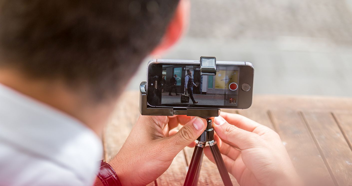 Ein Mann fotografiert an einem Morgen in London mit seinem Smartphone, das auf einem Stativ steht, das auf einem Holztisch platziert ist. Dazu die Bildunterschrift: Für eine professionelle Wirkung ist die Stabilisierung beim Dreh unerlässlich. © Getty Images/MatusDuda