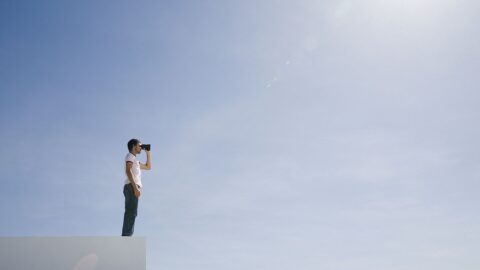 Ein Mann steht auf einem Beton-artigen Kasten und schaut durch ein Fernglas in die Ferne. Die Person steht vor einem klaren blauen Himmel. Dazu die Bildunterschrift: Distanz haben zu dem, über das man berichtet: eine Grundlage journalistischen Arbeitens. © Martin Barraud/Getty Images