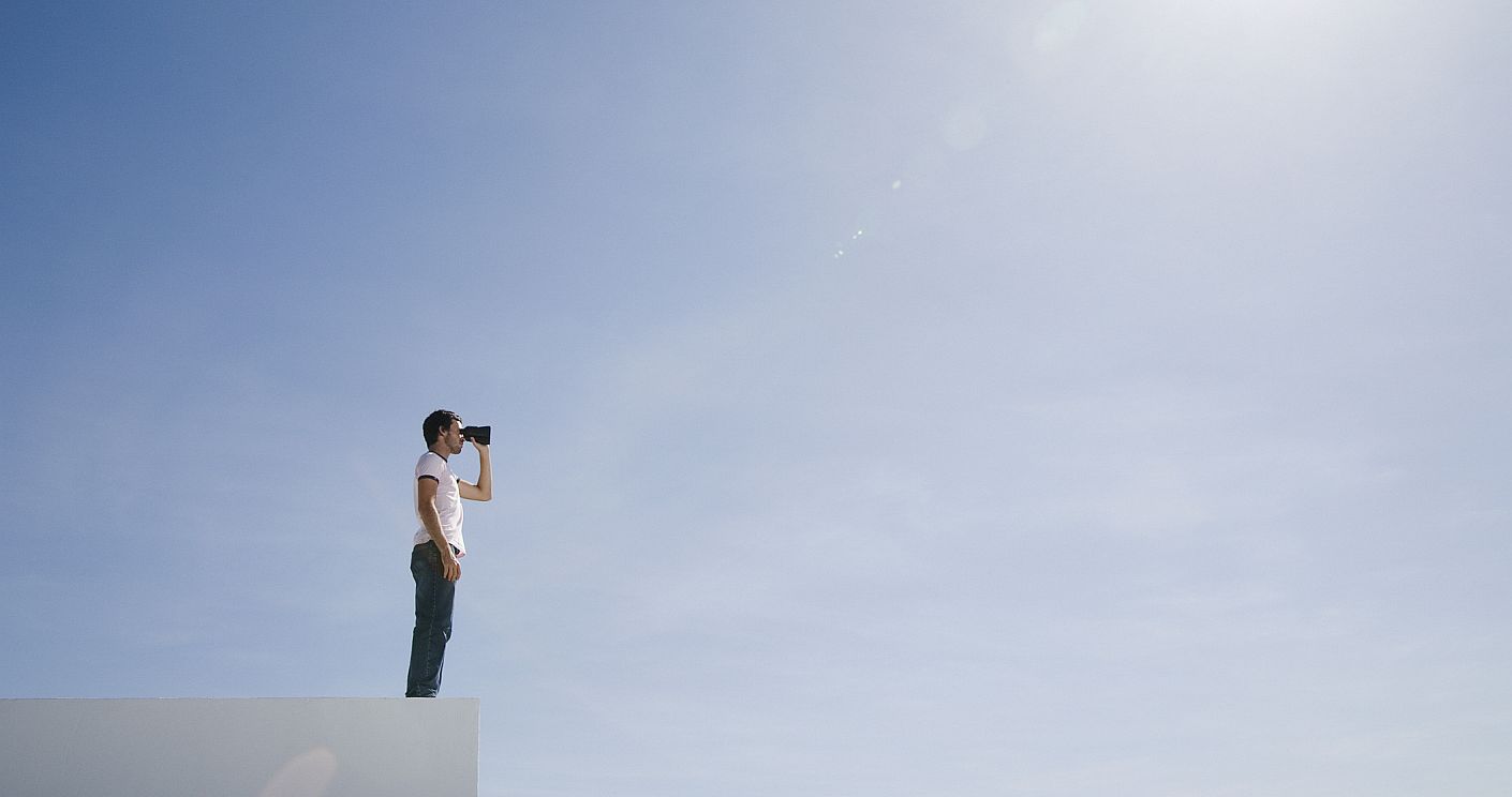 Ein Mann steht auf einem Beton-artigen Kasten und schaut durch ein Fernglas in die Ferne. Die Person steht vor einem klaren blauen Himmel. Dazu die Bildunterschrift: Distanz haben zu dem, über das man berichtet: eine Grundlage journalistischen Arbeitens. © Martin Barraud/Getty Images