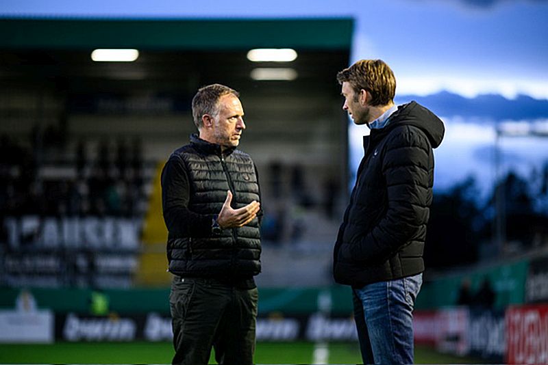 Zwei Männer im Gespräch: Links steht Holger Tromp, der die Kommunikation von Bayer Leverkusen leitet. Er spricht mit Simon Rolfes, Geschäftsführer Sport. Im Hintergrund ist ein Ausschnitt der leeren Stadiontribüne vor dunkelblauem und bewölkten Abendhimmel zu sehen. © privat