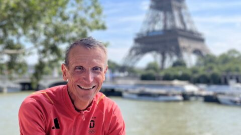 Adidas-Kommunikationschef Jan Runau in Trainingsjacke mit dem Schriftzug "Germany" lächelt in die Kamera. Im Hintergrund sind die Seine und der Eiffelturm zu sehen. Dazu die Bildunterschrift: Adidas-Kommunikationschef Jan Runau während der Olympischen Spiele 2024 in Paris. Der Besuch von Sportereignissen wie Olympia oder der Fußball-EM gehört für ihn zum Job. © privat