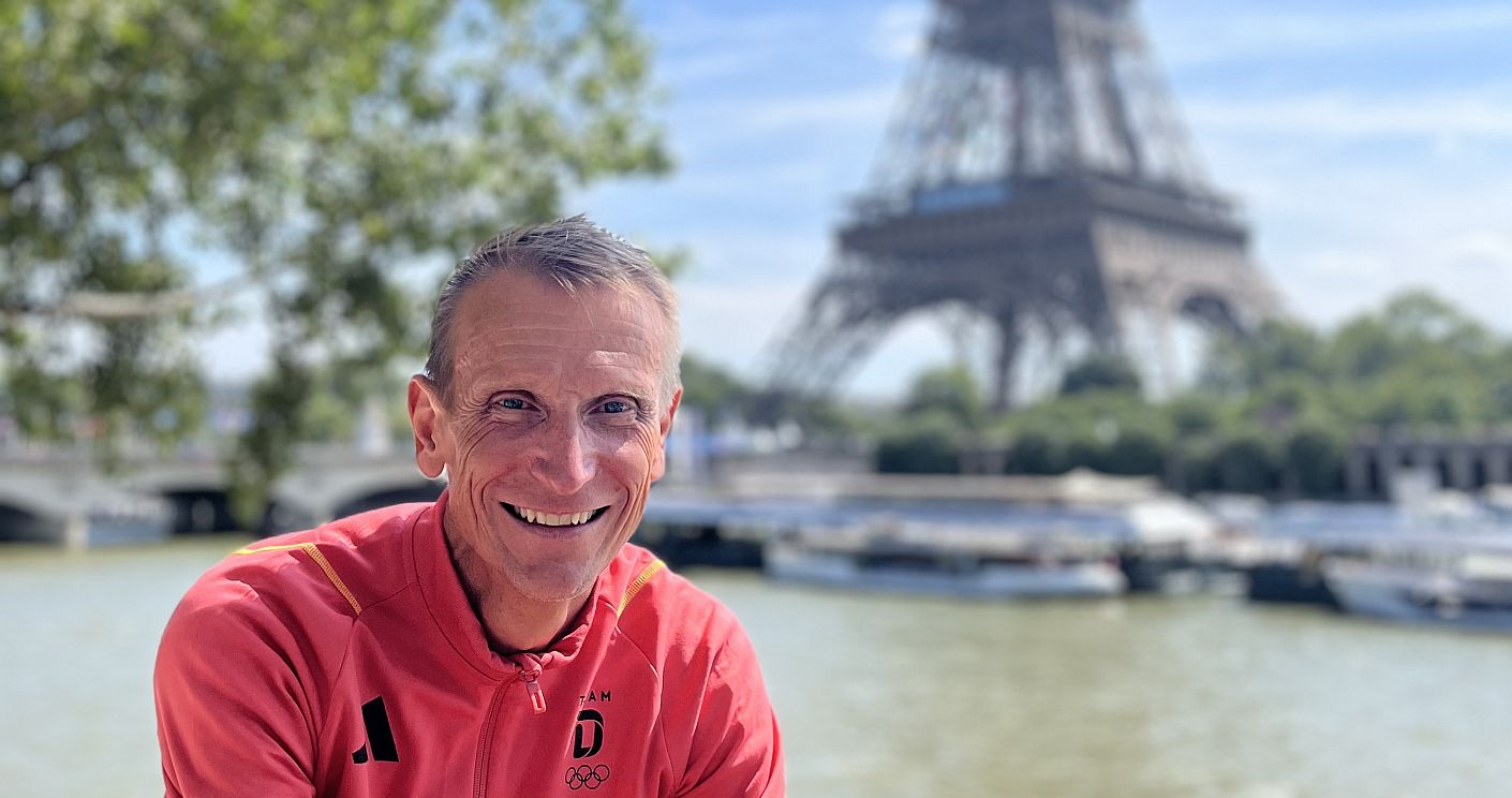 Adidas-Kommunikationschef Jan Runau in Trainingsjacke mit dem Schriftzug "Germany" lächelt in die Kamera. Im Hintergrund sind die Seine und der Eiffelturm zu sehen. Dazu die Bildunterschrift: Adidas-Kommunikationschef Jan Runau während der Olympischen Spiele 2024 in Paris. Der Besuch von Sportereignissen wie Olympia oder der Fußball-EM gehört für ihn zum Job. © privat