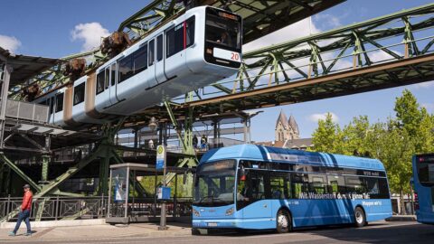 Ein blauer Wasserstoffbus hält an einer Haltestelle in Wuppertal. Über dem Bus fährt die Schwebebahn aus einem Bahnhof heraus. Dazu die Bildunterschrift: In Wuppertal fahren wasserstoffbetriebene Linienbusse. © Stefan Tesche-Hasenbach