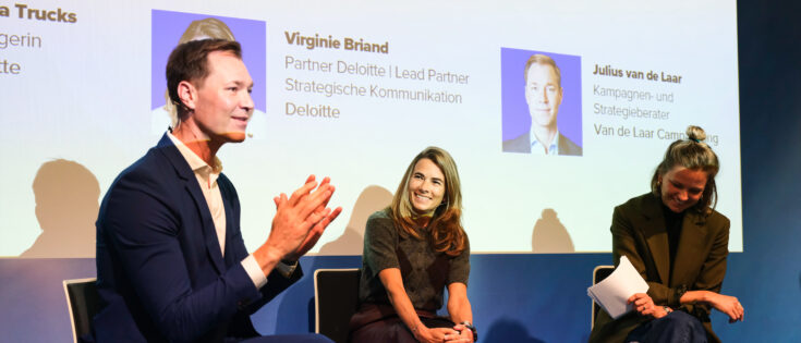 Drei Personen sitzen auf einer Bühne und diskutieren, im Hintergrund ein Bildschirm mit dem Titel des Beitrags.