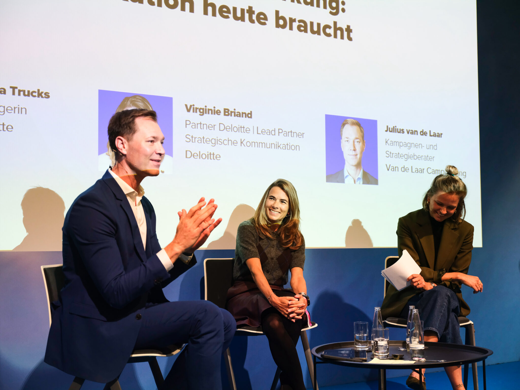 Drei Personen sitzen auf einer Bühne und diskutieren, im Hintergrund ein Bildschirm mit dem Titel des Beitrags.