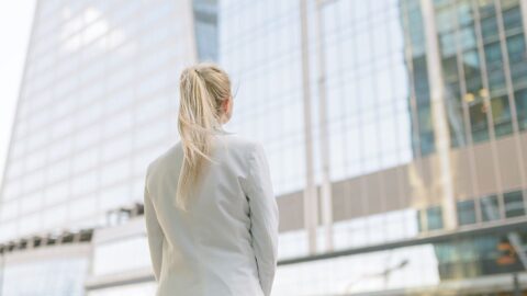 Zu sehen ist von hinten eine junge Frau mit weißem Blazer, die vor einem Bürogebäude steht. Dazu die Bildunterschrift: Nur ein Drittel der 40 Dax-Kommunikationschefs ist weiblich. Ein Grund: Es gibt wenige vakante Stellen. © Getty Images/Arndt_Vladimir