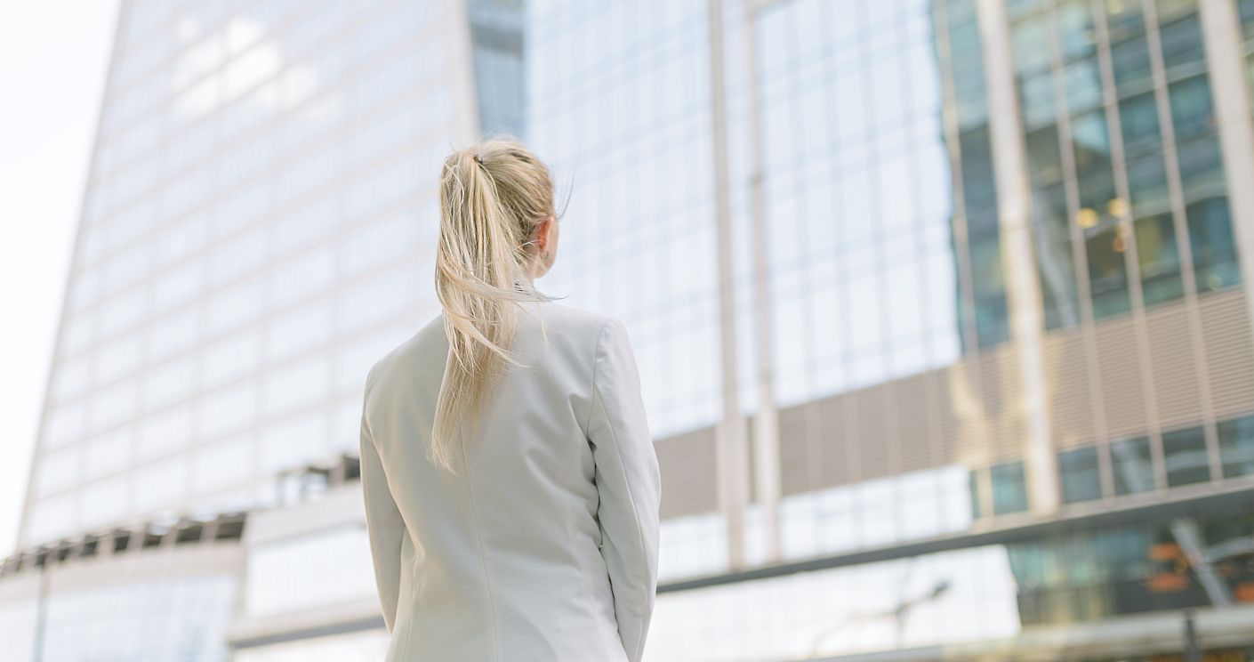 Zu sehen ist von hinten eine junge Frau mit weißem Blazer, die vor einem Bürogebäude steht. Dazu die Bildunterschrift: Nur ein Drittel der 40 Dax-Kommunikationschefs ist weiblich. Ein Grund: Es gibt wenige vakante Stellen. © Getty Images/Arndt_Vladimir