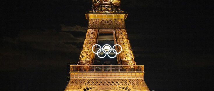 Der beleuchtete Eiffelturm während der Olympischen Spiele im Sommer 2024 in Paris (Frankreich). Am Turm sind erleuchtete Olympischen Ringe angebracht, hindurch leuchtet ein Vollmond. Dazu die Bildunterschrift: Die Olympischen Spiele in Paris lieferten starke Bild-Momente. Einmal stand der Mond so, dass er durch die Ringe am Eiffelturm schien. © IOC