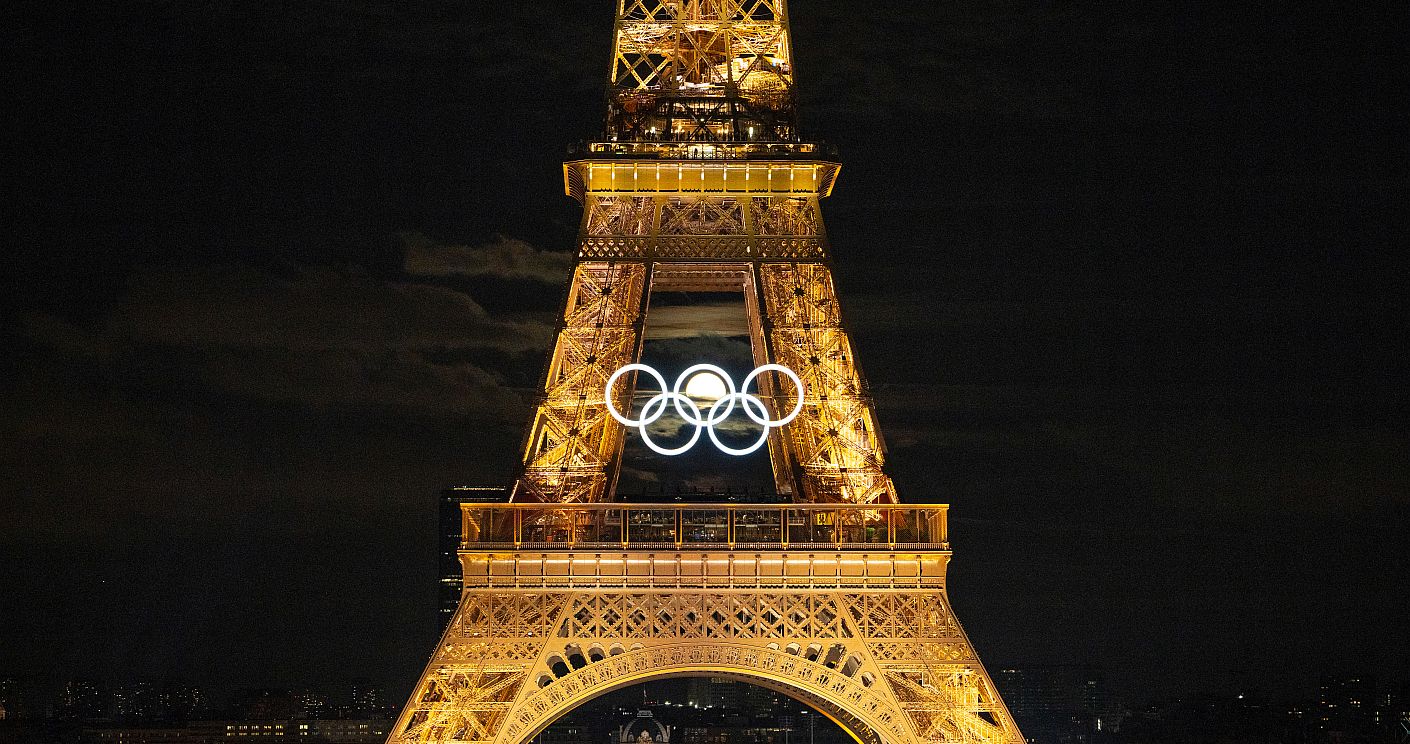 Der beleuchtete Eiffelturm während der Olympischen Spiele im Sommer 2024 in Paris (Frankreich). Am Turm sind erleuchtete Olympischen Ringe angebracht, hindurch leuchtet ein Vollmond. Dazu die Bildunterschrift: Die Olympischen Spiele in Paris lieferten starke Bild-Momente. Einmal stand der Mond so, dass er durch die Ringe am Eiffelturm schien. © IOC
