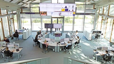 Ein sehr großer, lichtdurchfluteter Raum mit bodentiefen Fensterfronten. Menschen arbeiten vor Computerbildschirmen. In jeder Ecke stehen Schreibtischinseln. Im Zentrum eine Schreibtischfront in U-Form. An der vordersten Fensterfront sind übergroße Bildschirme angebracht mit Daten. Dazu die Bildunterschrift: Der Newsroom der Lufthansa ist darauf ausgerichtet, schnell reagieren zu können. © Lufthansa Group; Oliver Roesler