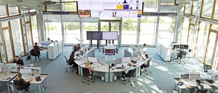 Ein sehr großer, lichtdurchfluteter Raum mit bodentiefen Fensterfronten. Menschen arbeiten vor Computerbildschirmen. In jeder Ecke stehen Schreibtischinseln. Im Zentrum eine Schreibtischfront in U-Form. An der vordersten Fensterfront sind übergroße Bildschirme angebracht mit Daten. Dazu die Bildunterschrift: Der Newsroom der Lufthansa ist darauf ausgerichtet, schnell reagieren zu können. © Lufthansa Group; Oliver Roesler