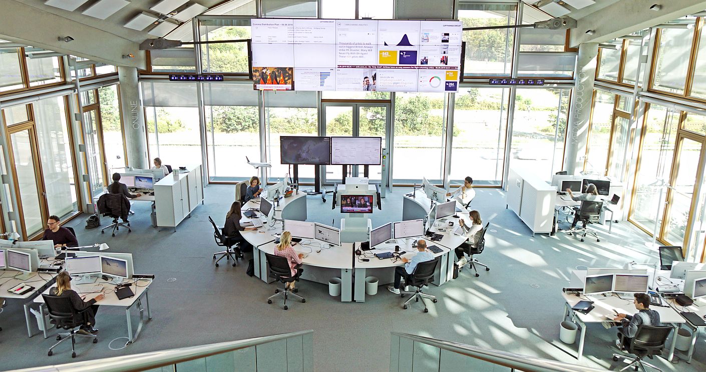 Ein sehr großer, lichtdurchfluteter Raum mit bodentiefen Fensterfronten. Menschen arbeiten vor Computerbildschirmen. In jeder Ecke stehen Schreibtischinseln. Im Zentrum eine Schreibtischfront in U-Form. An der vordersten Fensterfront sind übergroße Bildschirme angebracht mit Daten. Dazu die Bildunterschrift: Der Newsroom der Lufthansa ist darauf ausgerichtet, schnell reagieren zu können. © Lufthansa Group; Oliver Roesler
