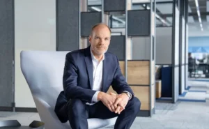 WAGNER_P_0074 | KOM - Magazin für Kommunikation Mann in Anzug sitzt entspannt in einem modernen Büro, umgeben von minimalistischer Einrichtung.