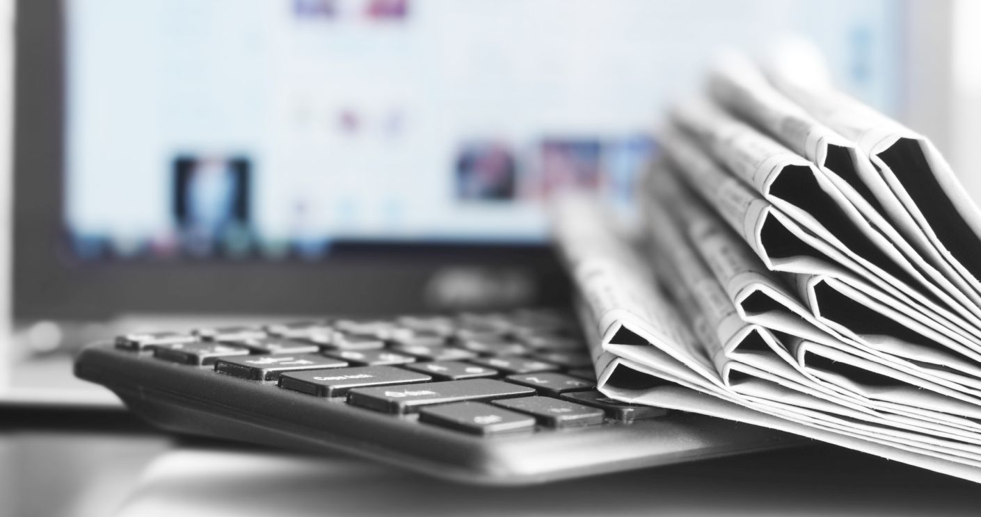 Laptop mit Tastatur im Vordergrund, daneben aufgeschichtete Zeitungen, im Hintergrund unscharfer Bildschirm. Dazu die Bildunterschrift: Journalisten informieren sich über Pressemitteilungen und persönliche Kontakte. © Getty Images/Fedor Kozyr
