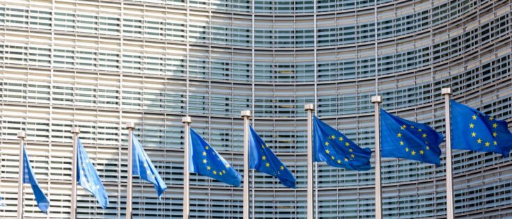 Reihe europäischer Flaggen weht im Wind vor dem Berlaymont-Gebäude der Europäischen Kommission in Brüssel. Dazu die Bildunterschrift: Wesentliche Entscheidungen zur ESG-Regulatorik werden auf EU-Ebene getroffen. © Olivier Rateau / Getty Images