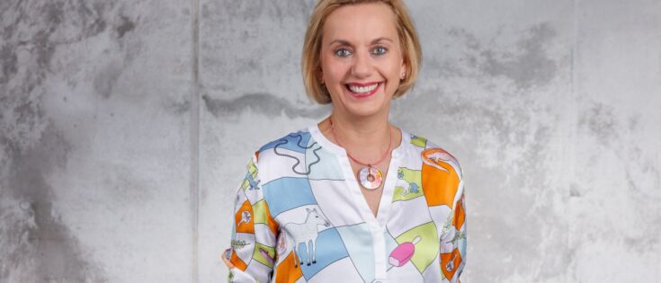 Frau mit blonden Haaren in bunter Bluse steht vor einer grauen Wand und lächelt freundlich. Dazu die Bildunterschrift: Alles unter einem Dach: Susanne Marell verantwortet bei der Schwarz Gruppe Kommunikation, Nachhaltigkeit, Public Affairs und Marke. © Matthias Stark