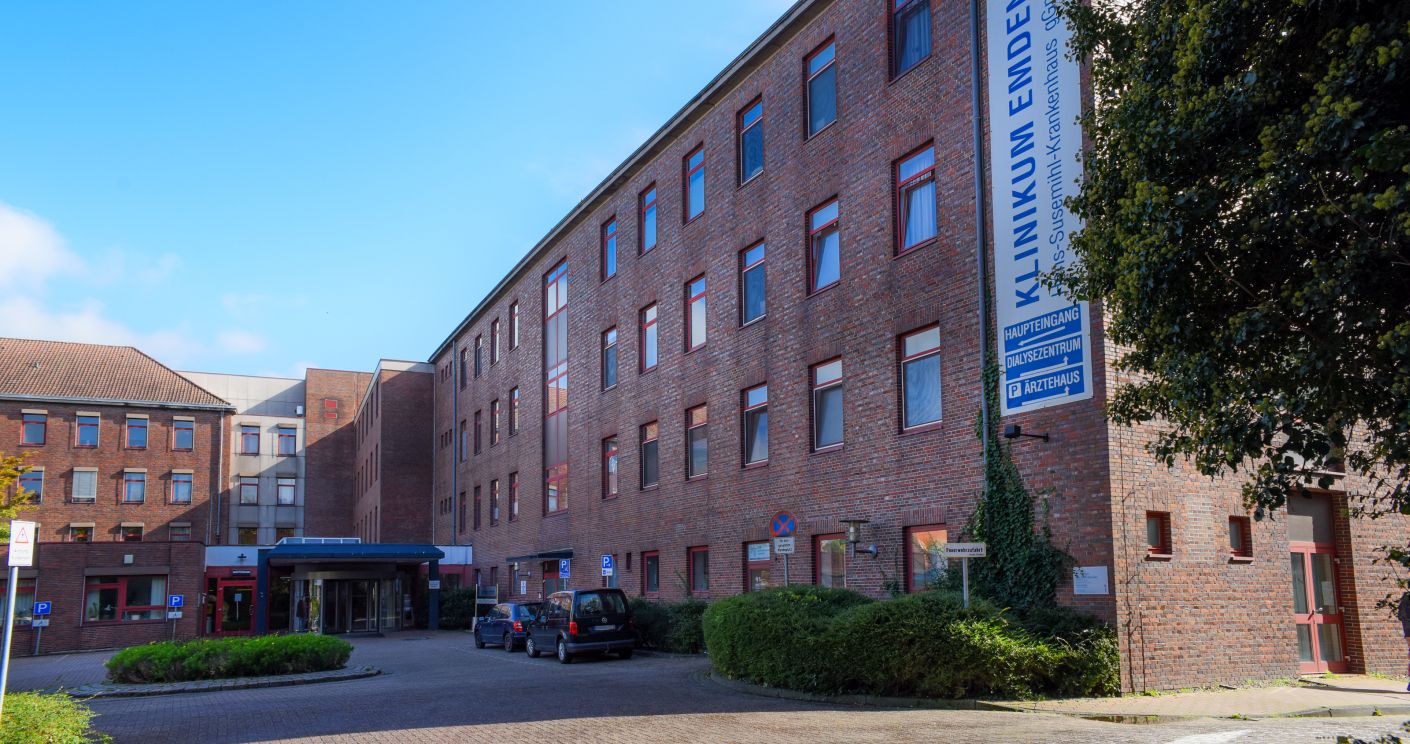 Klinikum Emden: Mehrstöckiges Backsteingebäude mit Eingangsbereich, Bäumen und parkenden Autos.