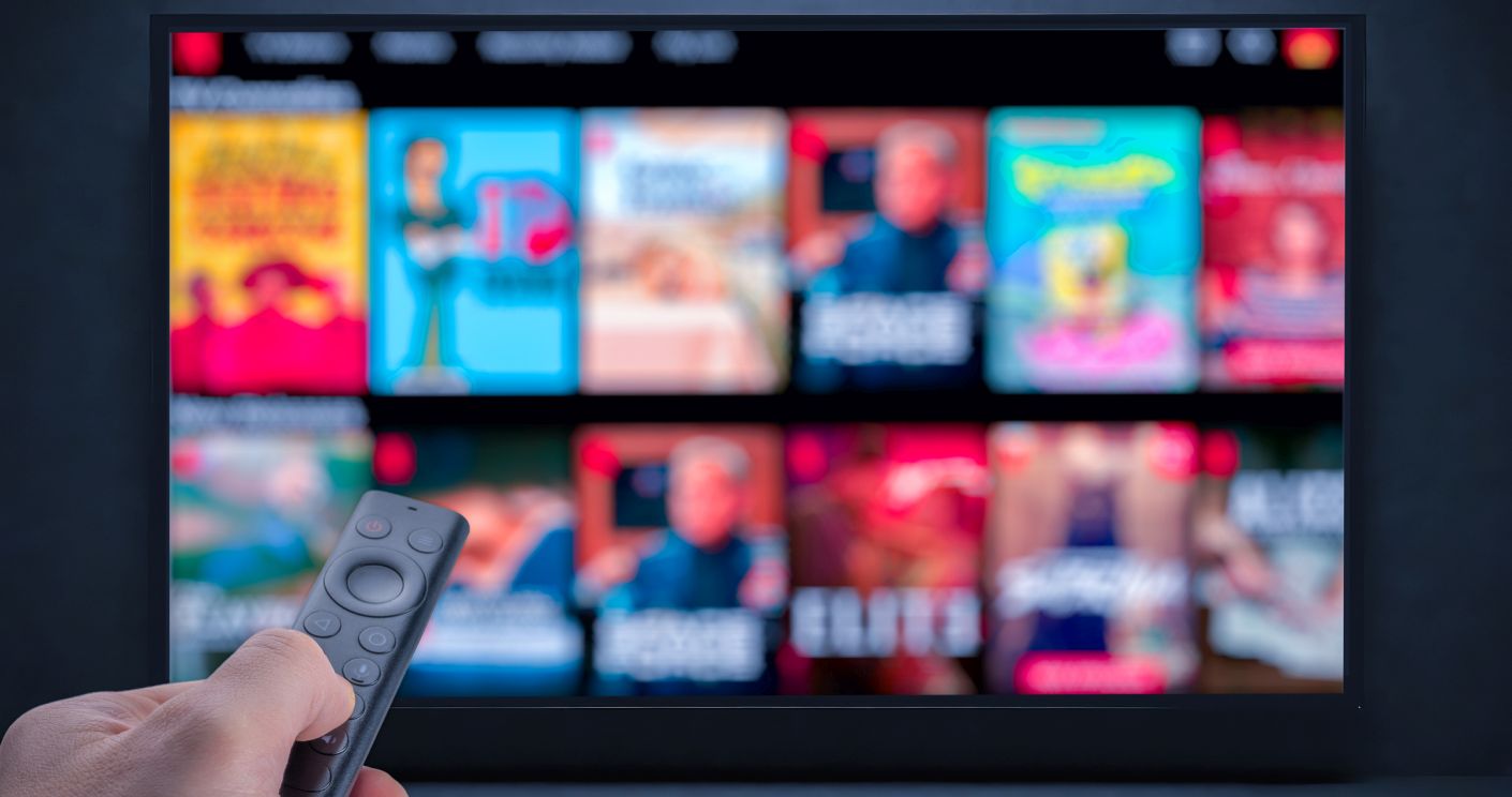 Eine Hand hält eine TV-Fernbedienung vor einem Bildschirm, auf dem verschiedene Streaming-Inhalte und Serien zu sehen sind.